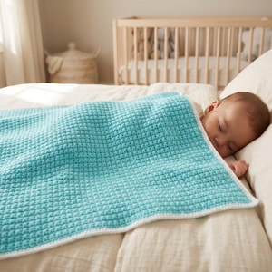 Pode incluir: Um bebé a dormir está aninhado sob uma manta turquesa de croché com uma borda branca. A manta tem um padrão texturizado, semelhante a um waffle. Um berço é visível no fundo, sugerindo um ambiente de berçário. A cena é banhada por uma luz suave e natural.