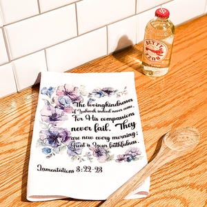 Op de afbeelding: Een witte keukendoek met een bloemenontwerp en de tekst "The lovingkindnesses of Yahweh indeed never cease...Great is Your faithfulness." Een houten lepel en een fles Betty Buzz zijn ook aanwezig.