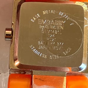 Pu&ograve; includere: Primo piano di un orologio da polso con cinturino arancione e cassa in metallo color argento. Il quadrante dell'orologio non &egrave; visibile. Il retro dell'orologio riporta la scritta "EMBASSY by GRUEN" e "STAINLESS STEEL BACK".