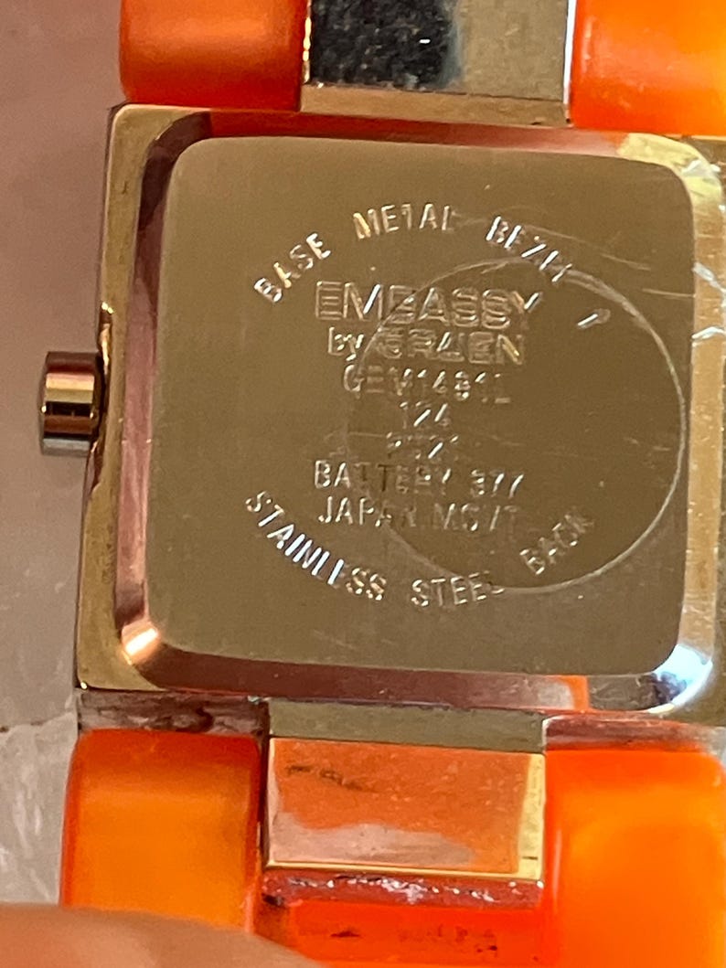 Pu&ograve; includere: Primo piano di un orologio da polso con cinturino arancione e cassa in metallo color argento. Il quadrante dell'orologio non &egrave; visibile. Il retro dell'orologio riporta la scritta "EMBASSY by GRUEN" e "STAINLESS STEEL BACK".