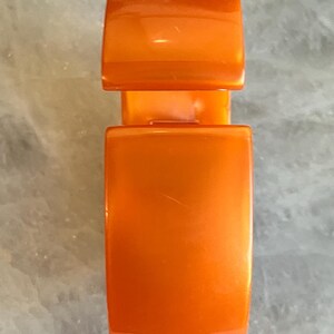 Pu&ograve; includere: Chiusura arancione lucida, di forma rettangolare con bordi arrotondati. La chiusura &egrave; composta da due parti: una sezione superiore pi&ugrave; piccola e una sezione inferiore pi&ugrave; grande, collegate da un ponte. Lo sfondo &egrave; una superficie chiara e strutturata.