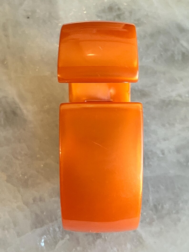 Pu&ograve; includere: Chiusura arancione lucida, di forma rettangolare con bordi arrotondati. La chiusura &egrave; composta da due parti: una sezione superiore pi&ugrave; piccola e una sezione inferiore pi&ugrave; grande, collegate da un ponte. Lo sfondo &egrave; una superficie chiara e strutturata.
