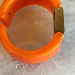Pu&ograve; includere: Bracciale rigido arancione brillante con cerniera. Il bracciale ha una forma arrotondata e una chiusura rettangolare color oro. Il bracciale &egrave; realizzato in materiale lucido su sfondo chiaro.