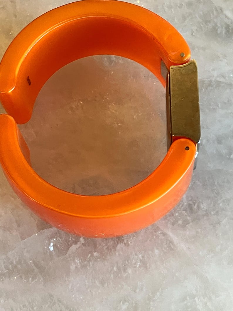 Pu&ograve; includere: Bracciale rigido arancione brillante con cerniera. Il bracciale ha una forma arrotondata e una chiusura rettangolare color oro. Il bracciale &egrave; realizzato in materiale lucido su sfondo chiaro.