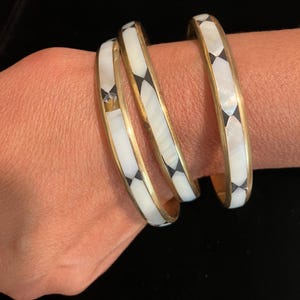 Może przedstawiać: Trzy okrągłe bransoletki typu bangle ze złotą metalową ramką, inkrustowane naprzemiennymi segmentami z białej masy perłowej oraz małymi czarnymi akcentami w kształcie rombów. Bransoletki mają gładkie, polerowane wykończenie i wzór.
