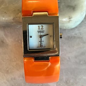 Pu&ograve; includere: Orologio da polso arancione con quadrante quadrato color argento. Il quadrante &egrave; azzurro con lancette nere e il numero 12. Il testo "EMBASSY by GRUEN" &egrave; stampato sul quadrante. Il cinturino &egrave; arancione lucido.