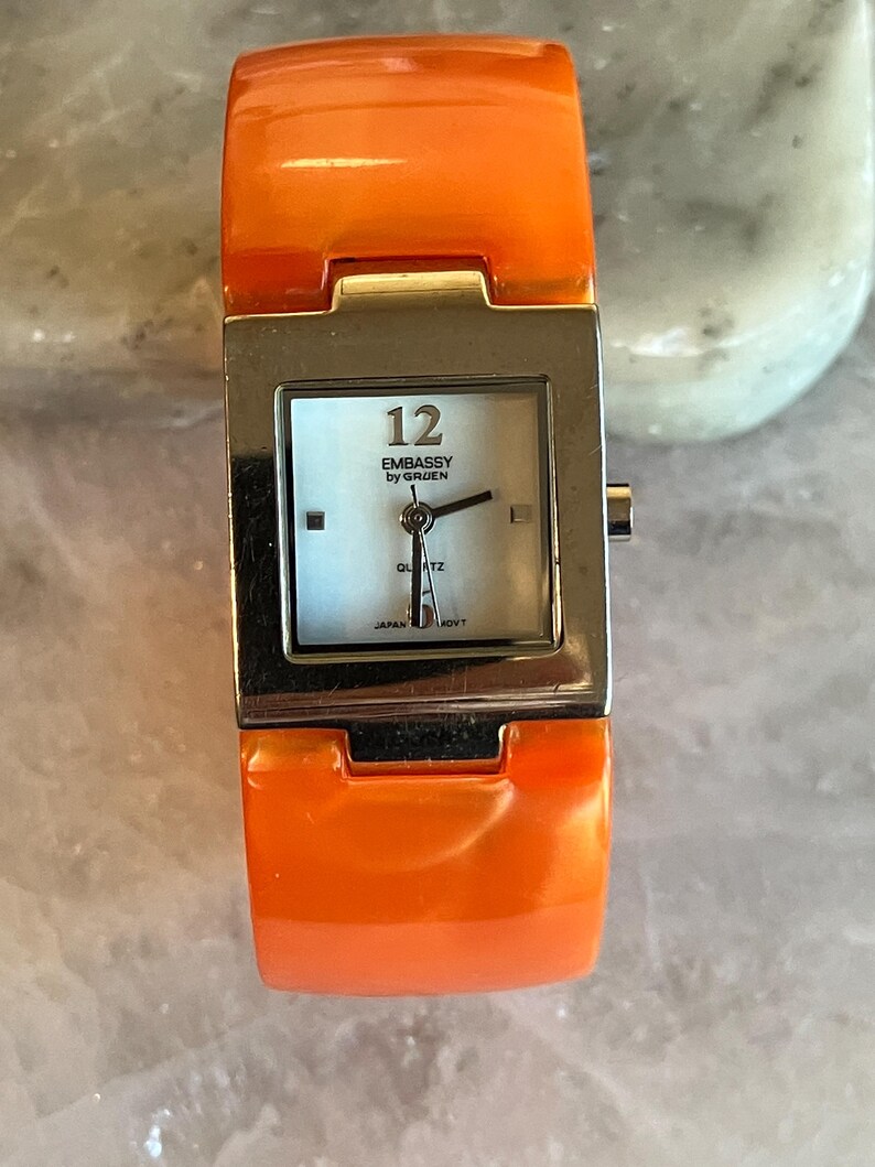 Pu&ograve; includere: Orologio da polso arancione con quadrante quadrato color argento. Il quadrante &egrave; azzurro con lancette nere e il numero 12. Il testo "EMBASSY by GRUEN" &egrave; stampato sul quadrante. Il cinturino &egrave; arancione lucido.