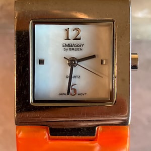 Pu&ograve; includere: Un orologio da polso quadrato con cassa color argento e cinturino arancione. Il quadrante &egrave; bianco con lancette e numeri neri, incluso il numero 12. L'orologio &egrave; marchiato "EMBASSY by GRUEN" e "JAPAN MOVT".