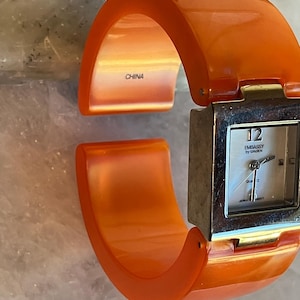 Pu&ograve; includere: Orologio da polso arancione con quadrante quadrato color argento. Il quadrante ha uno sfondo bianco con numeri e lancette nere. L'orologio &egrave; marchiato "Embassy by Groen" e "Japan Movt". L'interno del bracciale &egrave; marchiato "China".
