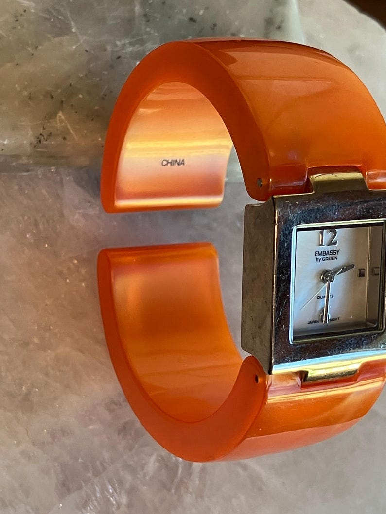 Pu&ograve; includere: Orologio da polso arancione con quadrante quadrato color argento. Il quadrante ha uno sfondo bianco con numeri e lancette nere. L'orologio &egrave; marchiato "Embassy by Groen" e "Japan Movt". L'interno del bracciale &egrave; marchiato "China".