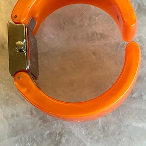 Pu&ograve; includere: Orologio a bracciale arancione brillante con quadrante rettangolare color argento. Il cinturino &egrave; spesso, curvo e arancione pieno. L'orologio &egrave; aperto, mostrando la chiusura e il quadrante.