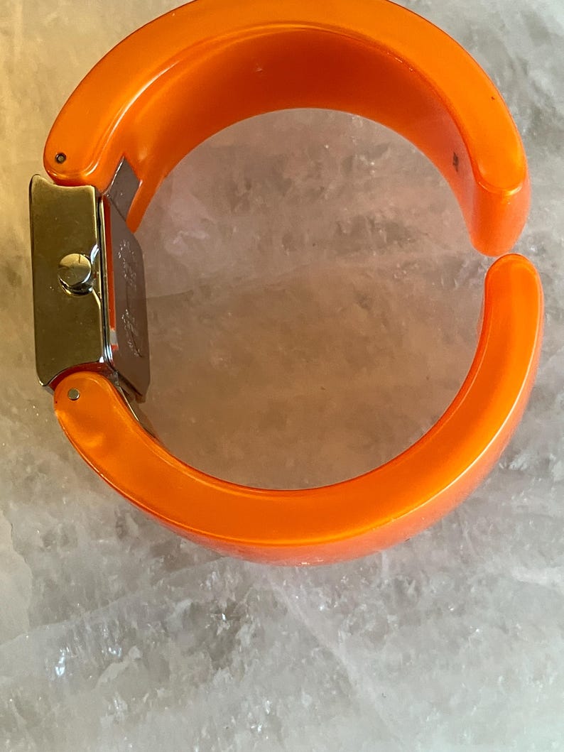 Pu&ograve; includere: Orologio a bracciale arancione brillante con quadrante rettangolare color argento. Il cinturino &egrave; spesso, curvo e arancione pieno. L'orologio &egrave; aperto, mostrando la chiusura e il quadrante.