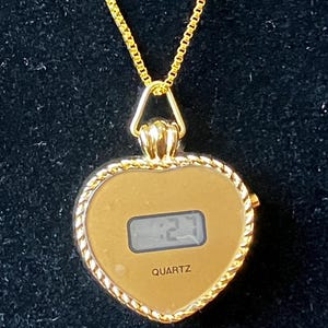 Puede incluir: Un colgante de reloj dorado en forma de corazón con una pantalla digital que muestra la hora. El reloj está suspendido de una delicada cadena dorada. La palabra "QUARTZ" está impresa debajo de la pantalla.
