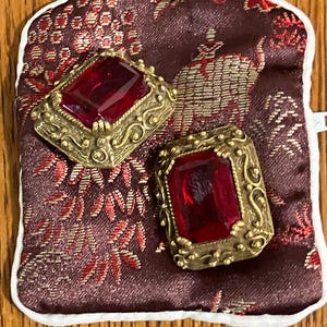 May include: A pair of vintage gold-toned earrings with large, rectangular, red gemstones. The earrings are set in ornate frames and rest on a burgundy fabric pouch with a floral pattern. The pouch has a white border and a white button closure.