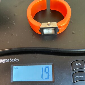 Pu&ograve; includere: Un orologio a bracciale arancione con un quadrante rettangolare color argento. L'orologio &egrave; su una bilancia che indica 54 grammi. La bilancia &egrave; nera e riporta la scritta "amazon basics".