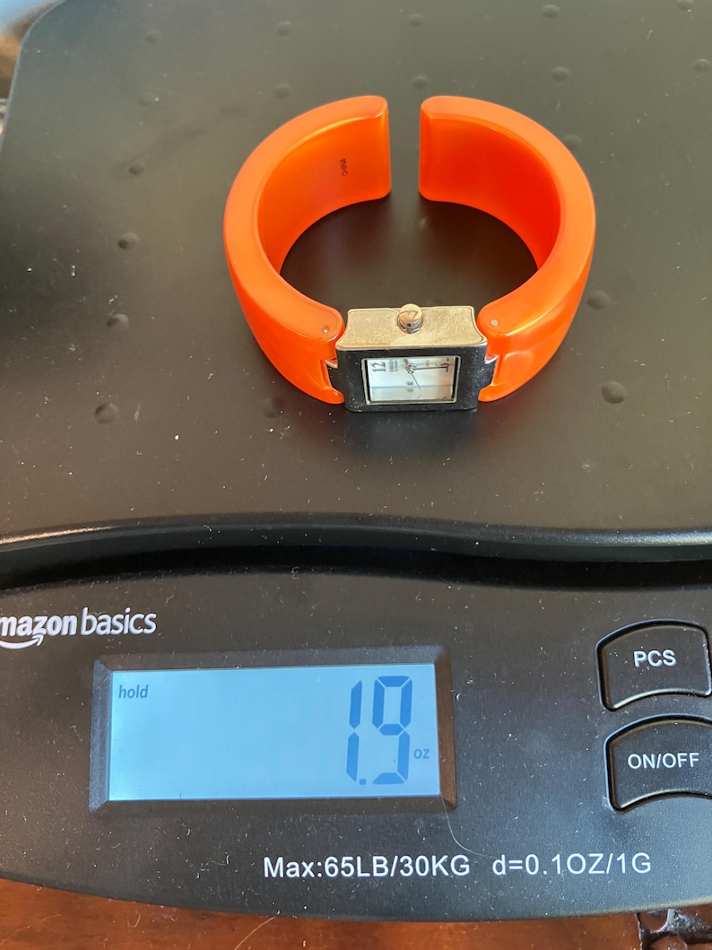 Pu&ograve; includere: Un orologio a bracciale arancione con un quadrante rettangolare color argento. L'orologio &egrave; su una bilancia che indica 54 grammi. La bilancia &egrave; nera e riporta la scritta "amazon basics".