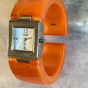 Pu&ograve; includere: Orologio da polso arancione con quadrante quadrato color argento. Il quadrante dell'orologio ha uno sfondo bianco madreperla con numeri e lancette nere. L'orologio &egrave; marchiato "EMBASSY by GRUEN" e "QUARTZ". L'orologio &egrave; marchiato "CHINA".