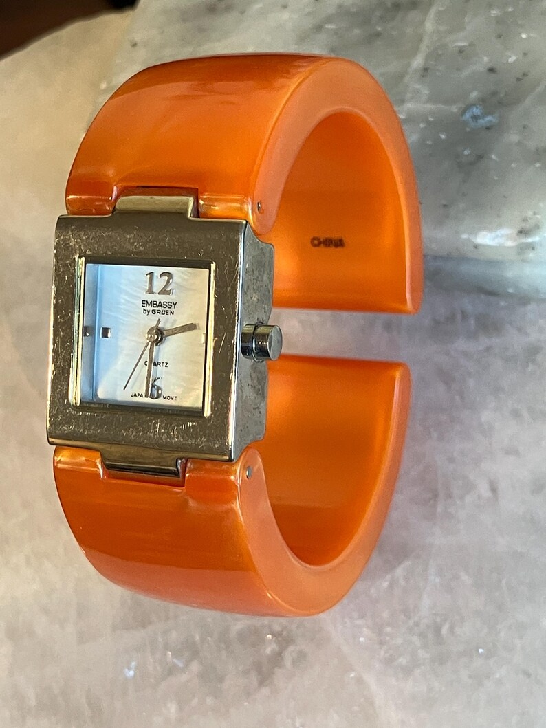 Pu&ograve; includere: Orologio da polso arancione con quadrante quadrato color argento. Il quadrante dell'orologio ha uno sfondo bianco madreperla con numeri e lancette nere. L'orologio &egrave; marchiato "EMBASSY by GRUEN" e "QUARTZ". L'orologio &egrave; marchiato "CHINA".