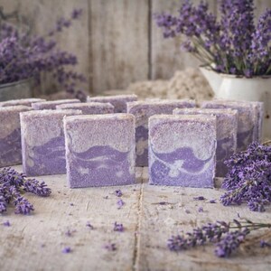 Puede incluir: Barras de jabón artesanal de lavanda con diseño veteado, dispuestas sobre una superficie de madera rústica. Ramos de lavanda fresca y una jarra vintage llena de ramitas de lavanda realzan la estética natural. Las barras de jabón son de color lila claro.