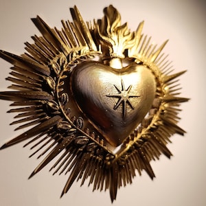 Può includere: Un emblema religioso a forma di cuore color oro con una stella al centro. Il cuore è circondato da raggi, fiamme e una corona d'alloro. L'emblema è probabilmente un oggetto devozionale o un'opera d'arte religiosa.