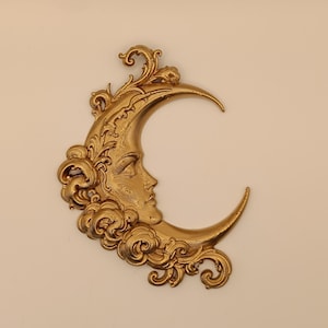 Può includere: Una decorazione da parete color oro a forma di mezzaluna con un volto umano di profilo. La luna è circondata da ornamenti a volute e motivi simili a nuvole che si estendono lungo il bordo esterno della forma a mezzaluna.