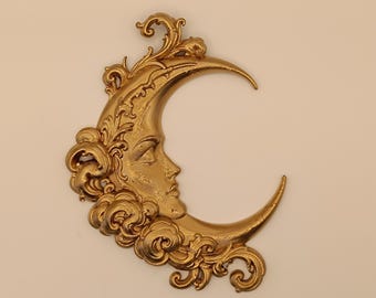 May include: A gold-colored wall decoration shaped like a crescent moon with a human face in profile. The moon is surrounded by ornate, swirling filigree and cloud-like patterns that extend along the outer edge of the crescent shape.