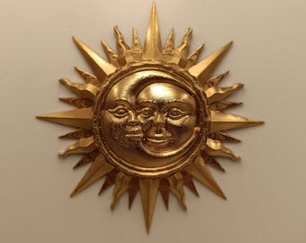 Könnte beinhalten: Ein goldfarbenes Wandornament mit einem zentralen kreisförmigen Design, das ein überlappendes Sonnen- und Mondgesicht zeigt. Das Stück ist von zahlreichen strahlenden, spitzen Strahlen unterschiedlicher Länge umgeben, die einen Sterneneffekt erzeugen.