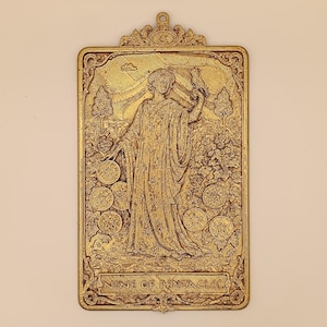 May include: A rectangular gold-toned metal wall plaque featuring a relief design of the Nine of Pentacles tarot card. The image depicts a figure in robes standing among vines with nine pentacles, a bird on their hand, and the text NINE OF PENTACLES.