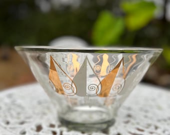 Puede incluir: Un cuenco de vidrio transparente con un diseño decorativo de hojas de oro y plata. El cuenco tiene una abertura ancha y una base redondeada. El diseño presenta formas de hojas doradas y espirales plateadas. El cuenco está sobre una mesa blanca ornamentada.