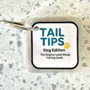 Puede incluir: Un tarjetero de plástico transparente con un anillo de metal, que contiene tarjetas de entrenamiento. Las tarjetas dicen "TAIL TIPS Dog Edition" en verde azulado, con una huella de pata amarilla. También se ve el texto "The Original Leash-Ready Training Cards".
