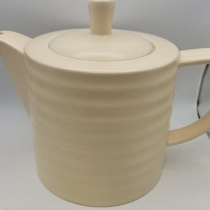 May include: A cream-colored ceramic teapot featuring a cylindrical body with horizontal ribbed detailing, a matching lid with a central knob, a curved spout, and a sturdy handle extending from the side of the vessel.