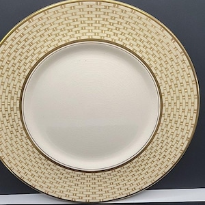 May include: A round, cream-colored plate with a woven basket pattern around the rim and a gold-colored border. The center of the plate is a solid cream color. The plate is a piece of dinnerware.