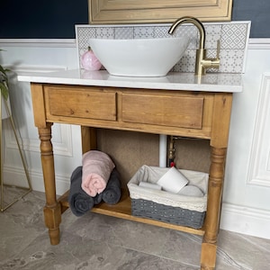 Peut inclure: Meuble-lavabo de salle de bain avec un plan de travail blanc, un lavabo vasque blanc et un robinet doré. La vanité a deux tiroirs et une étagère inférieure avec des serviettes roulées et un panier. Le cadre est en bois brun clair.