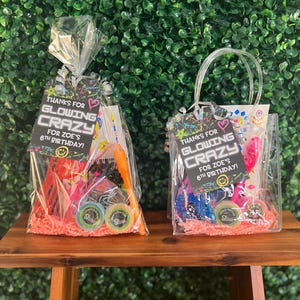 May include: Two clear gift bags filled with colorful toys and treats, each with a tag that reads "THANKS FOR GLOWING CRAZY FOR ZOE'S 6TH BIRTHDAY!". The bags sit on a small wooden table against a green hedge background.