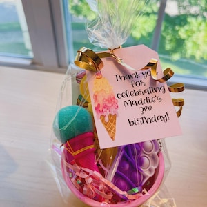 May include: A pink gift basket filled with toys and treats, wrapped in clear cellophane and tied with a gold ribbon. The basket includes a teal-colored toy, a pink cone-shaped item, and a purple pop-it toy. A pink tag reads "Thank you for celebrating Maddie's 3rd birthday!"