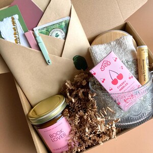 May include: A gift box filled with various items, including a candle labeled "Book Lover", a pink chocolate bar with cherry illustrations, a lip balm, and tea packets. The box also contains a small glass bowl, a washcloth, and stationery.