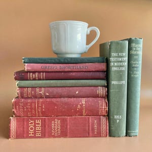 May include: A white ceramic mug sits atop a stack of vintage books. The stack includes titles such as Gregg Shorthand, Behind the Scenes, The New Testament in Modern English, Manna in the Morning, and a red Holy Bible Revised Standard Version.