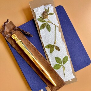 May include: A gold-toned metal pen rests on a brown suede-like pouch with a drawstring. Beside it is a rectangular bookmark made of white textured paper with deckled edges, featuring a pressed green clover plant with a small brown flower head.