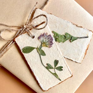 May include: Two rectangular off-white paper tags with deckled edges, each featuring a pressed dried flower. One tag displays a purple and white clover with green leaves, while the other shows a green sprig. Both are tied with brown twine.
