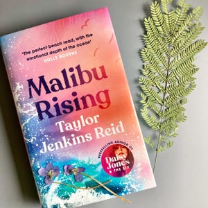 May include: A book titled Malibu Rising by Taylor Jenkins Reid with a pink, orange, and blue gradient cover featuring ocean waves, a circular badge for the author of Daisy Jones and the Six, and a quote by Holly Bourne, accented by two small purple flowers.