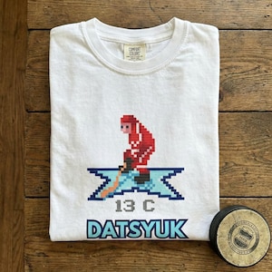 Könnte beinhalten: Weißes T-Shirt mit einer pixeligen Eishockeyspieler-Grafik in Rot und Blau, der Nummer "13 C" und dem Namen "Datsyuk" in Blau. Rechts ist ein Hockeypuck mit einem kreisförmigen Emblem.