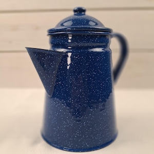 May include: A dark blue enamel coffee percolator featuring a white speckled pattern throughout the exterior. The vessel includes a matching lid with a small knob handle, a wide base, a tapered body, and a prominent triangular spout on the side.