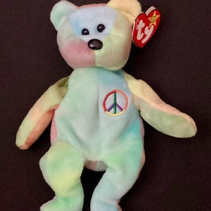May include: A colorful plush bear with a peace sign on its chest. The bear features a patchwork design in pastel shades of blue, green, pink, and yellow. It has a red heart-shaped tag attached to its ear.