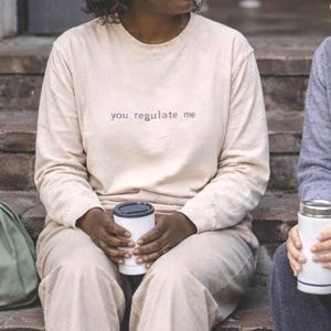 Op de afbeelding: Twee mensen zitten op bakstenen trappen, elk met een witte koffiekop. Eén draagt een beige sweatshirt met de tekst "you regulate me". De ander draagt een grijze hoodie. Een groene tas en een koffiekop zijn ook zichtbaar.