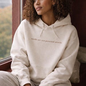 May include: Cream-coloured hoodie with the text "needs are not personal attacks" in a brown font. The hoodie has a drawstring hood and a front pocket. The model is wearing matching grey sweatpants.