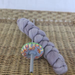 May include: A hand spindle with a mushroom design and a braid of gray wool. The spindle has a gray shaft, a mushroom-shaped top with a colorful design, and a brass hook. The wool braid is a soft, light gray color.