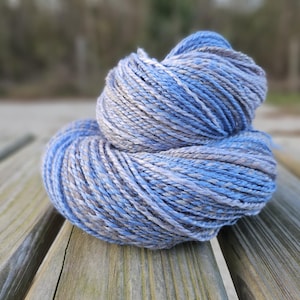 May include: A skein of yarn featuring a twisted, multi-ply construction with a variegated color palette of light blue, medium blue, and soft grey. The fibers are tightly wound into a rounded, textured bundle with a visible sheen throughout.