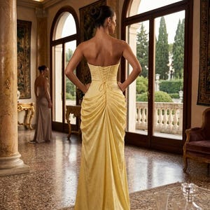 K&ouml;nnte beinhalten: Ein schulterfreies, bodenlanges gelbes Kleid mit Schn&uuml;rr&uuml;cken. Das Kleid hat ein drapiertes Design und wird von einer Person getragen, die in einem gro&szlig;en Raum mit gro&szlig;en Fenstern und aufwendiger Einrichtung steht. Eine andere Person ist im Hintergrund zu sehen, die ein &auml;hnliches Kleid tr&auml;gt.