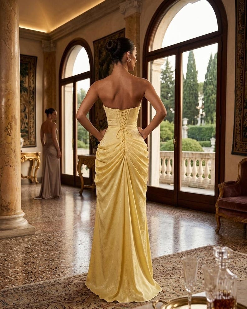 K&ouml;nnte beinhalten: Ein schulterfreies, bodenlanges gelbes Kleid mit Schn&uuml;rr&uuml;cken. Das Kleid hat ein drapiertes Design und wird von einer Person getragen, die in einem gro&szlig;en Raum mit gro&szlig;en Fenstern und aufwendiger Einrichtung steht. Eine andere Person ist im Hintergrund zu sehen, die ein &auml;hnliches Kleid tr&auml;gt.