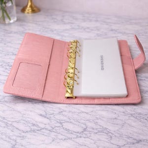 Op de afbeelding: Open roze leren binder met goudkleurige ringen en een kaartsleuf. De binder bevat wit papier met het woord "SPENDING" erop gedrukt. De binder ligt open op een marmeren oppervlak.